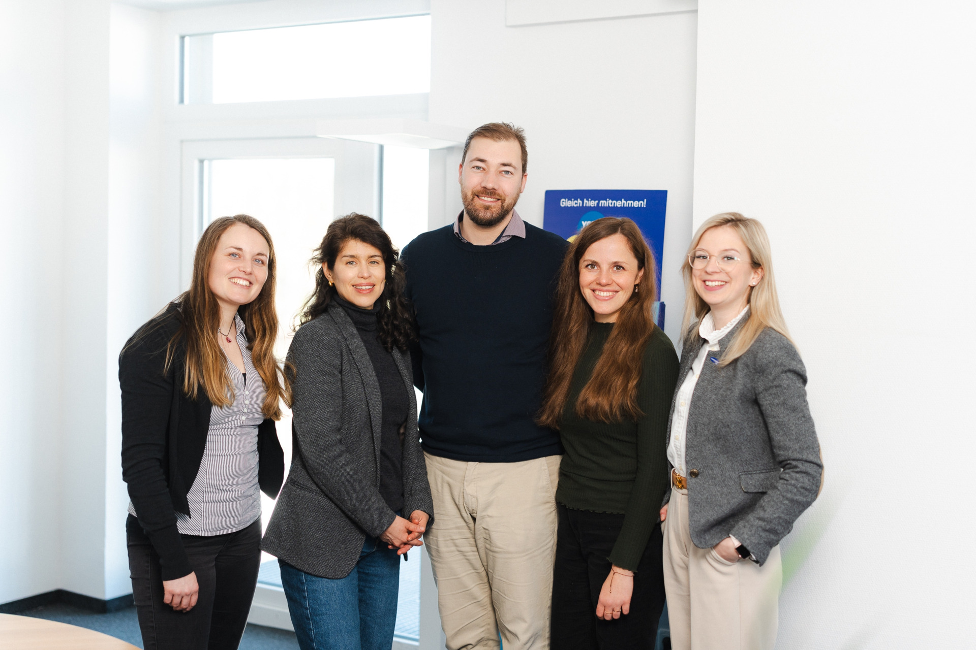  Group of five colleagues smiling in a modern office, showcasing teamwork and professional attire, against a bright background.