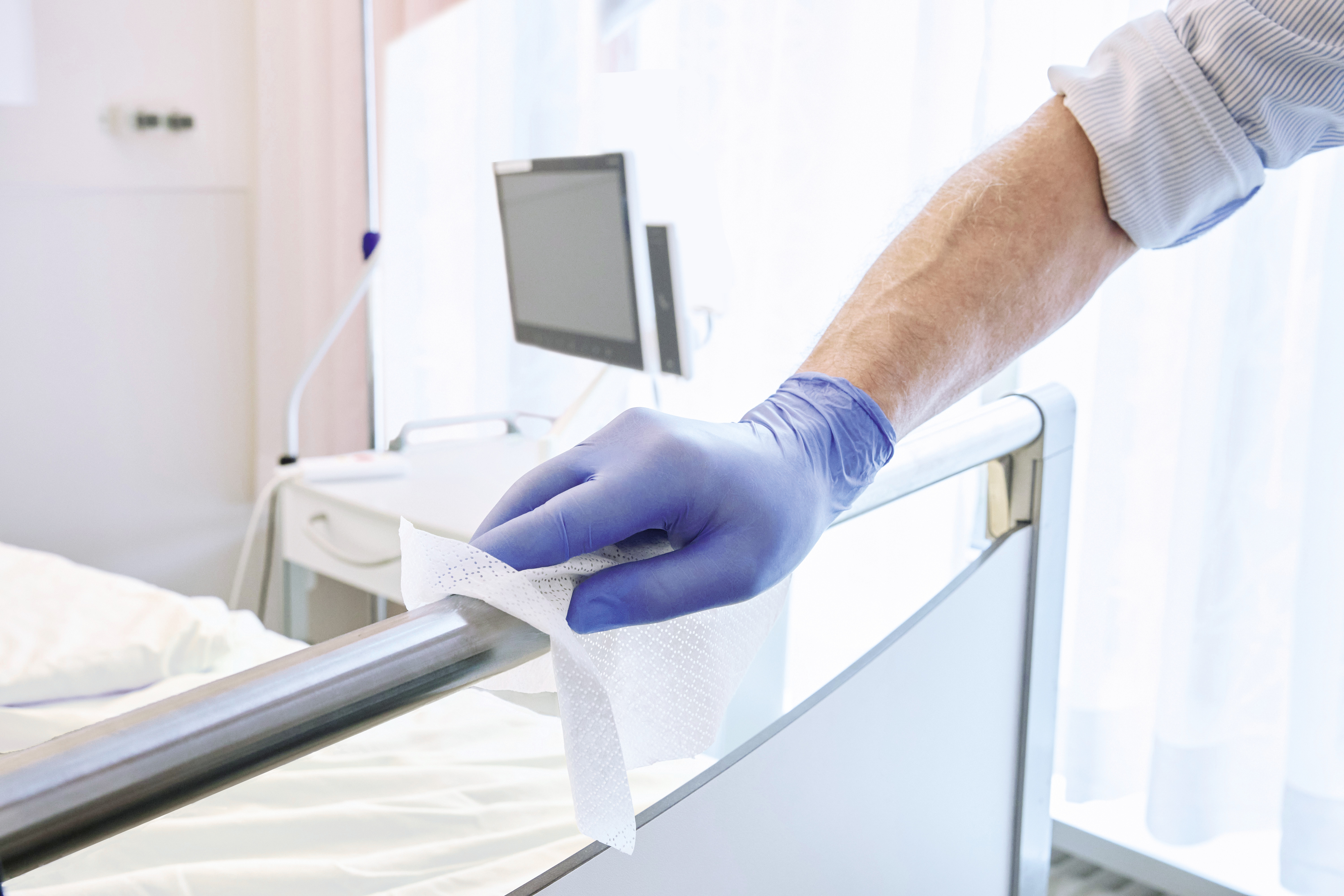 A person wearing a blue disposable glove disinfects a hospital bed rail with a wipe in a bright patient room.