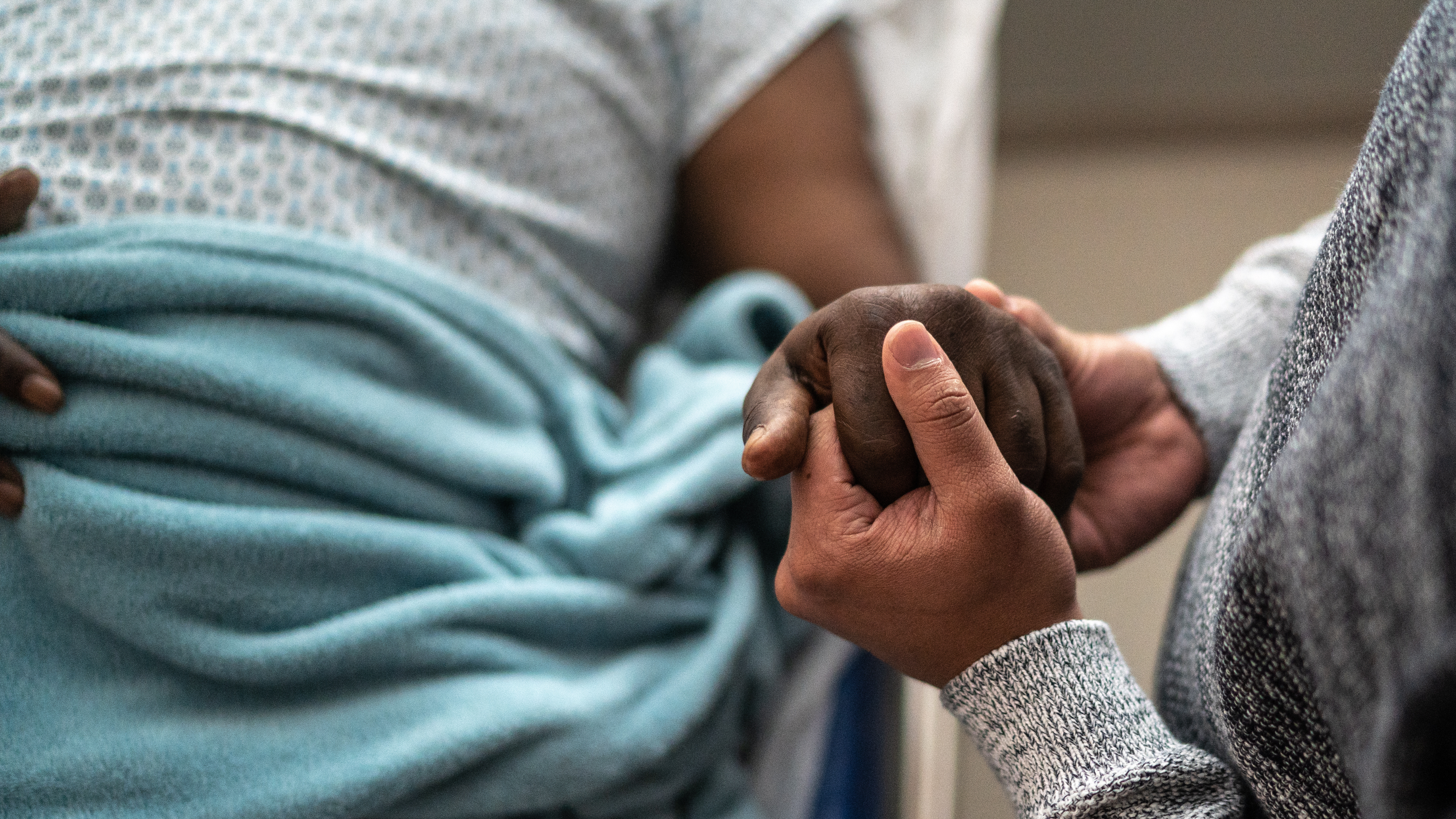 A caregiver gently holds a patient’s hand beside a hospital bed, conveying comfort and support.