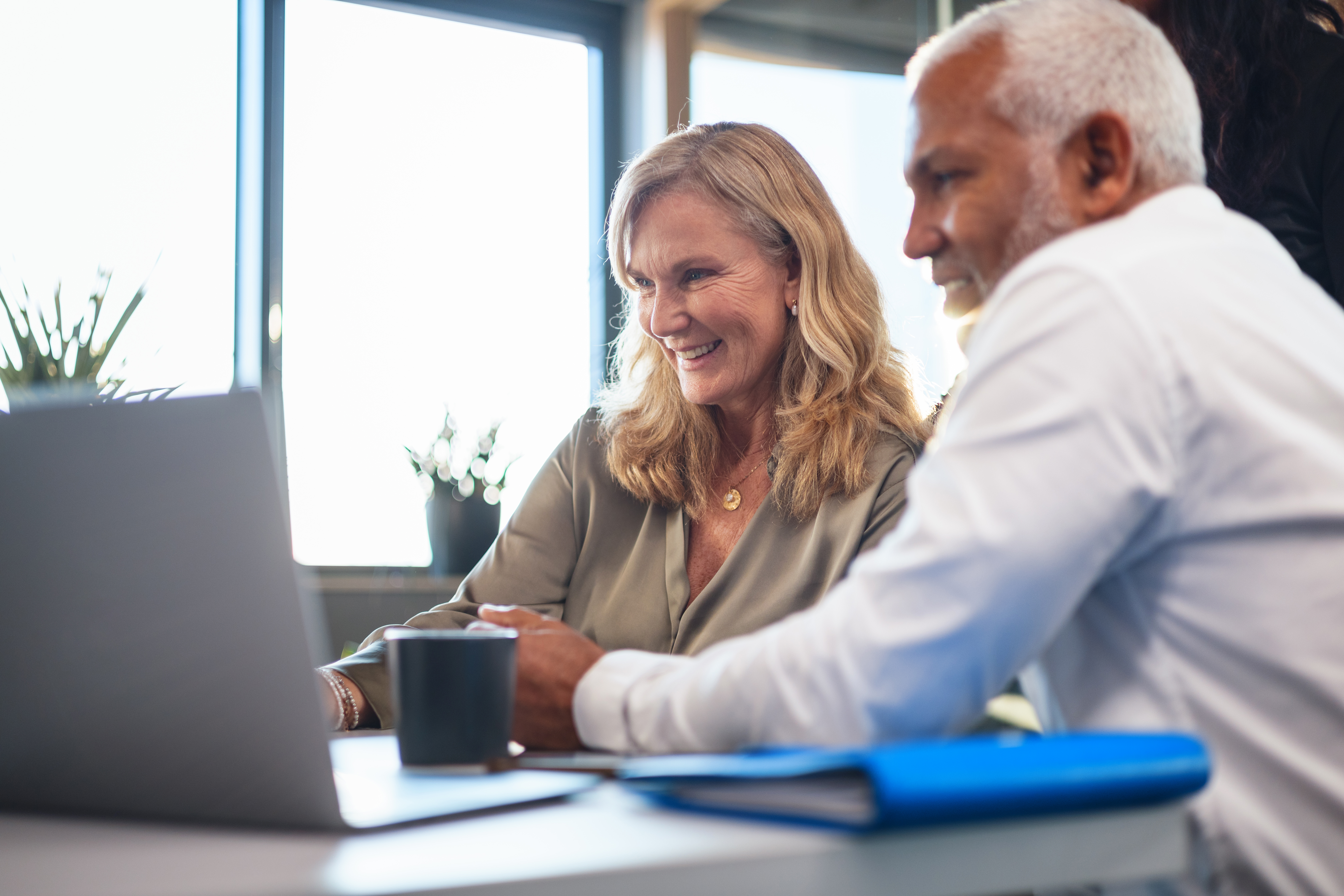  A middle-aged man and woman in a bright, modern office engage in collaborative work on a laptop. The atmosphere is professional and friendly, highlighting teamwork and diversity in the workplace.