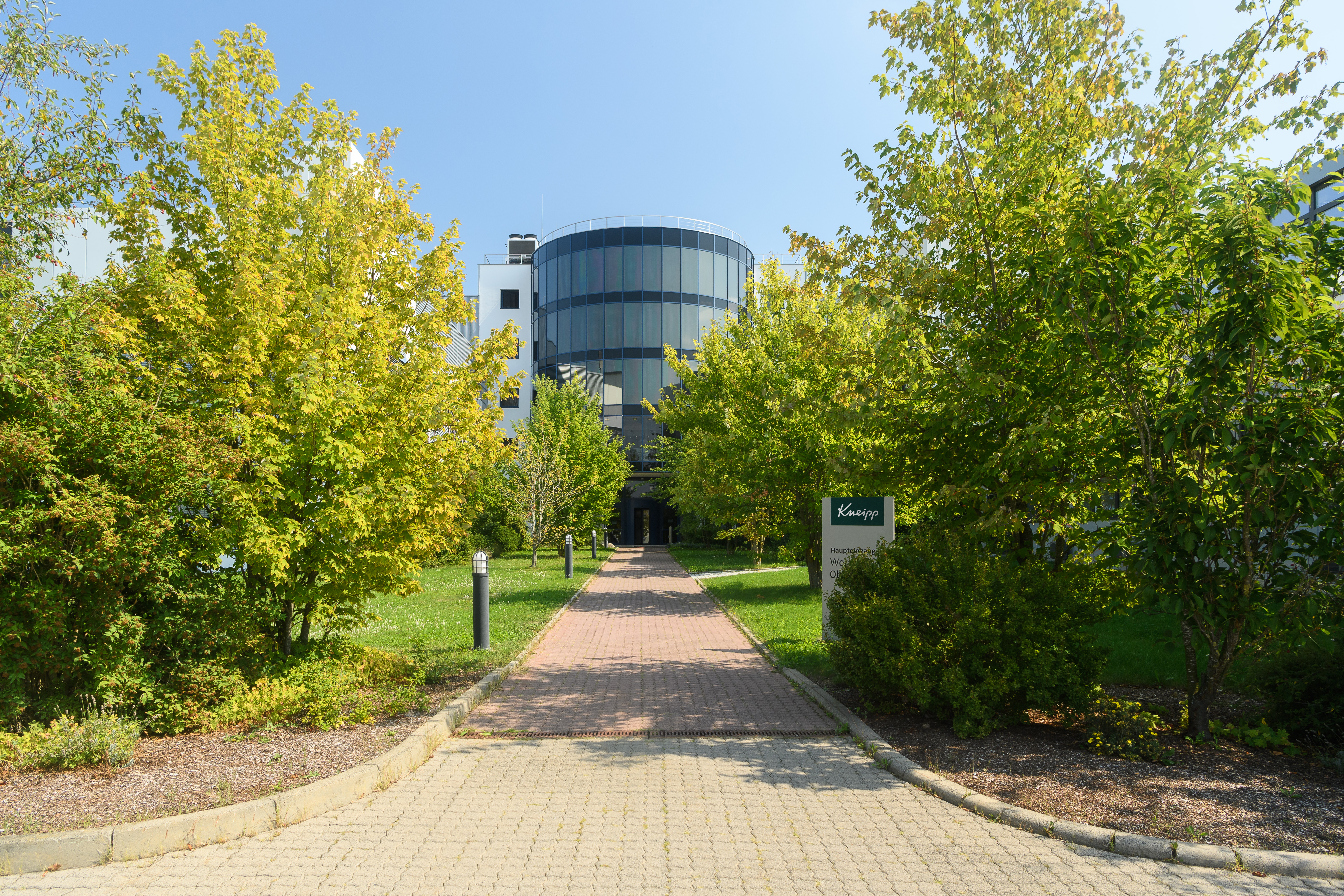  A modern office building with circular design surrounded by lush green trees and a neatly paved path leading to the entrance.