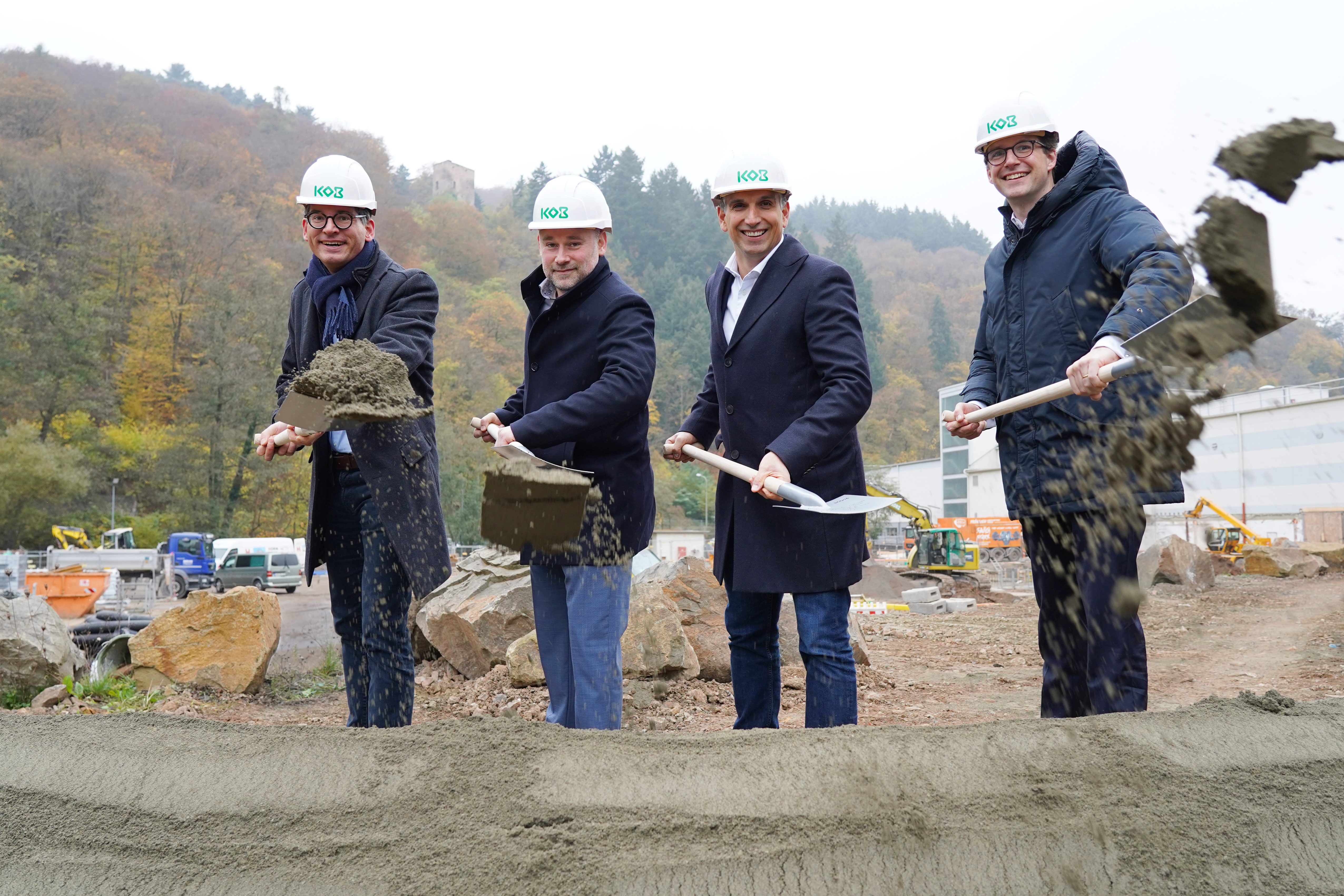 Four men in hard hats holding shovels, participating in a groundbreaking ceremony with a construction site and autumn trees in the background.