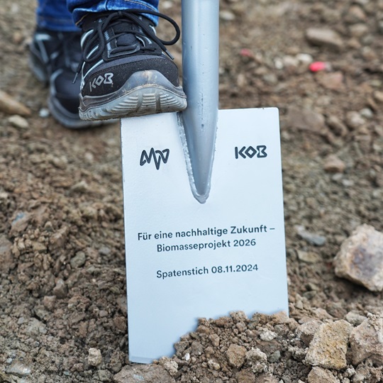 A foot wearing a black shoe steps on a spade with text: 'For a sustainable future – Biomass Project 2026. Groundbreaking 08.11.2024.'