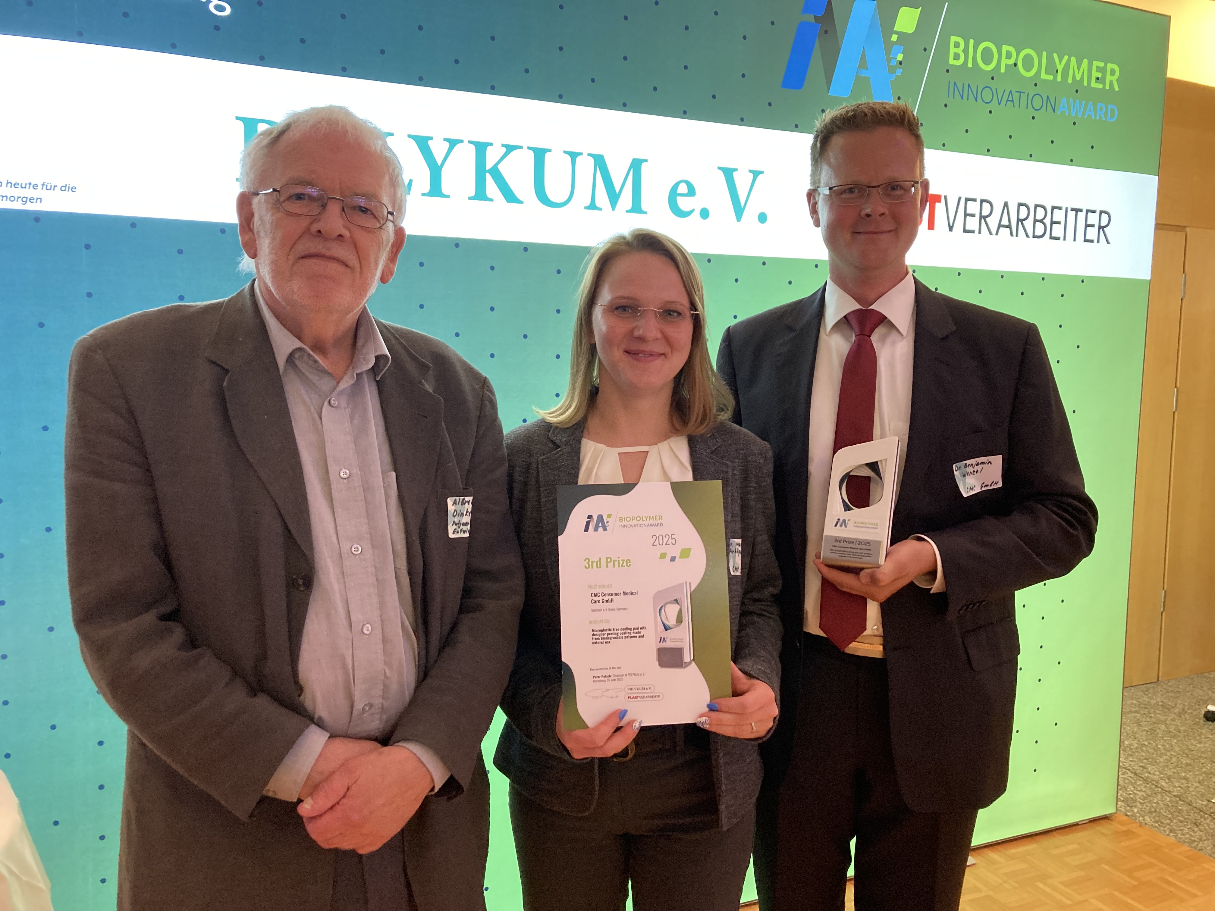  Three individuals at the Biopolymer Innovation Award ceremony, two holding certificates and a plaque, against a branded backdrop.