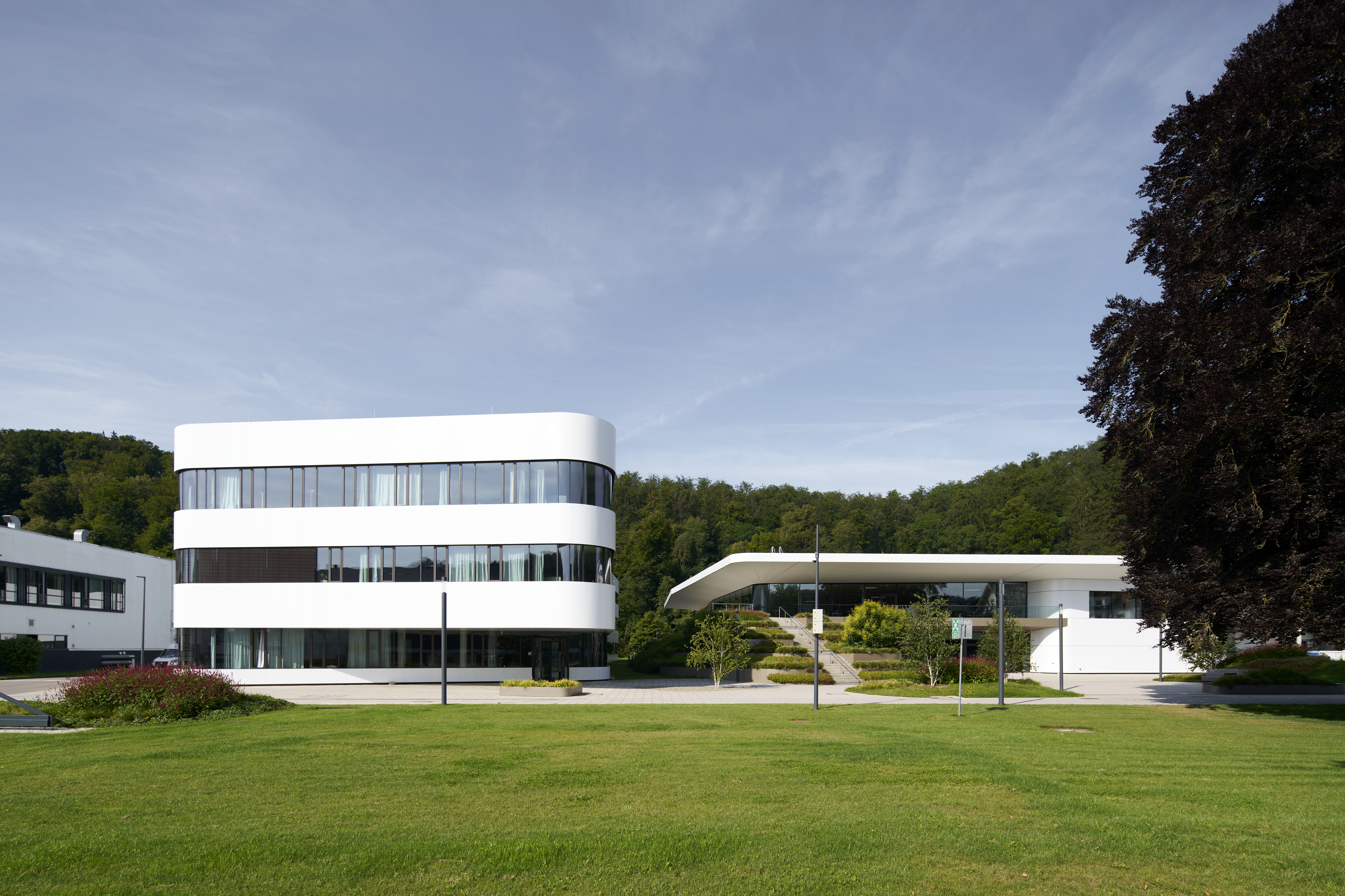 Contemporary white building with curved lines, surrounded by green lawns and trees, set against a clear blue sky.