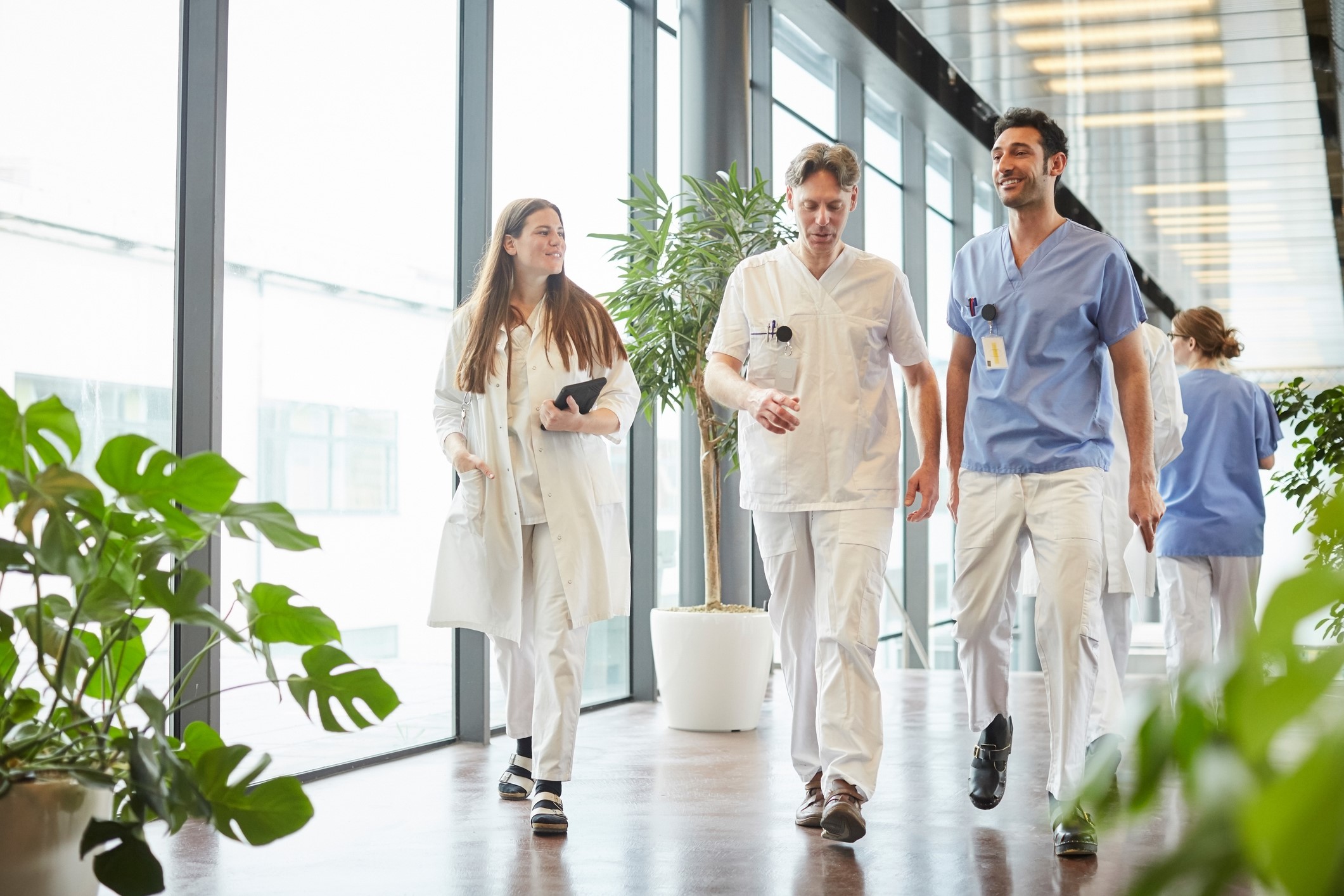  Group of three healthcare professionals walking in a bright medical facility corridor adorned with potted plants, exuding confidence and teamwork.